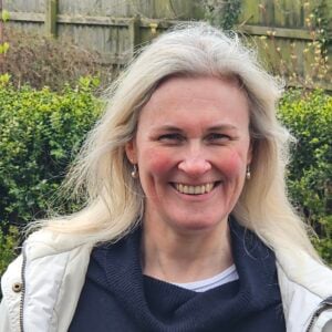 White woman with long, straight, blonde hair, wearing navy jumper and white coat, in front of green hedge.