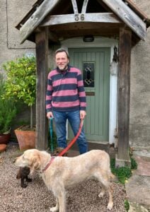 A man in a grey and pink stripey top and jeans, standing in front of a green front door. He is holding a golden retriever dog. A smaller brown dog can be partially seen behind the golden retriever. The man is Alan, the Green Party candidate for the Frampton Cotterell by-election. The dogs are not standing for election.