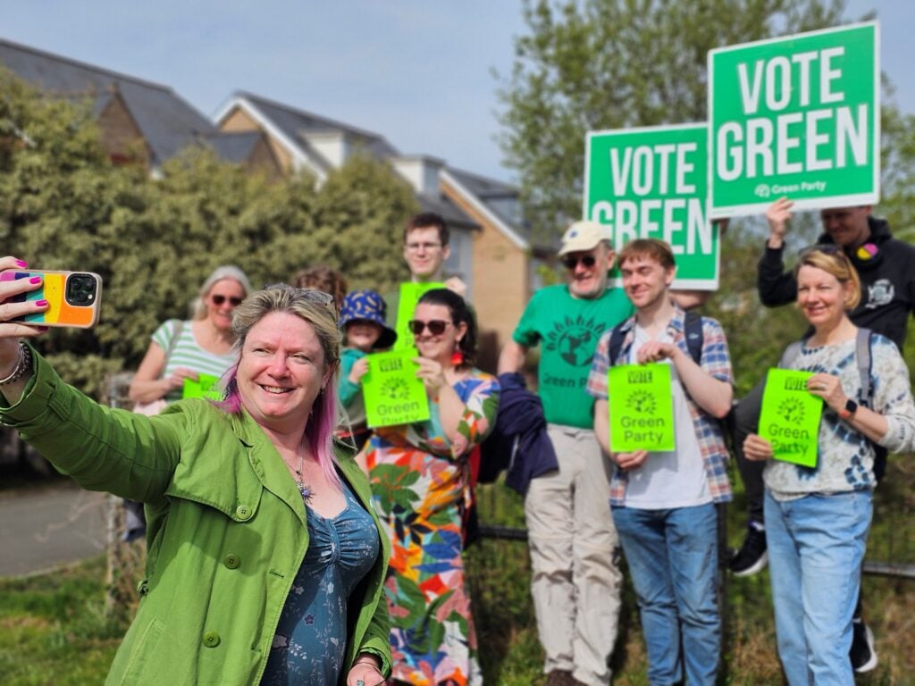 A white woman with blonde and pink hair, wearing green clothes, stands in front of a group of people who are holding up Vote Green signs.