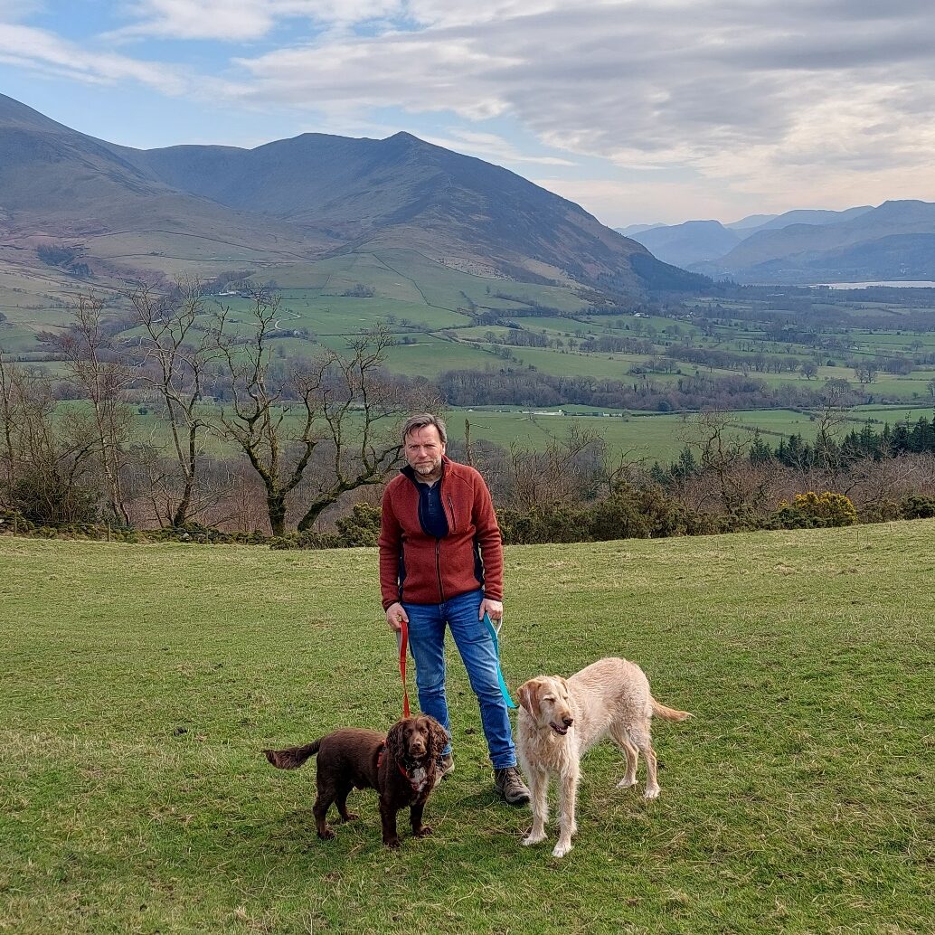 A man with two dogs, in a field, with hills in the background.